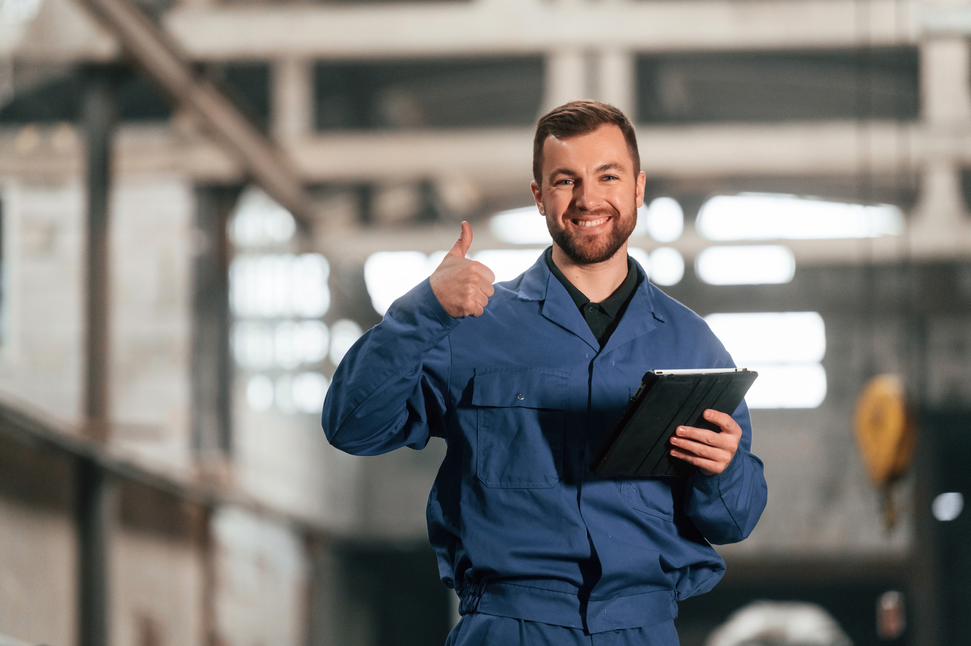 Smiling supply chain professional in blue overalls giving a thumbs up while holding a tablet